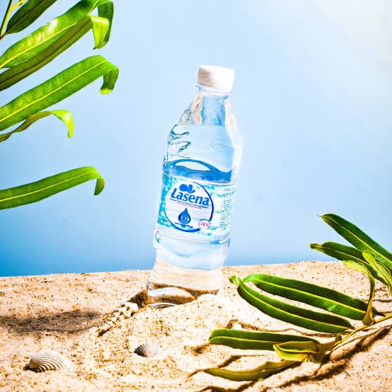 A Lasena bottle standing on sand beside tropical leaves and shells